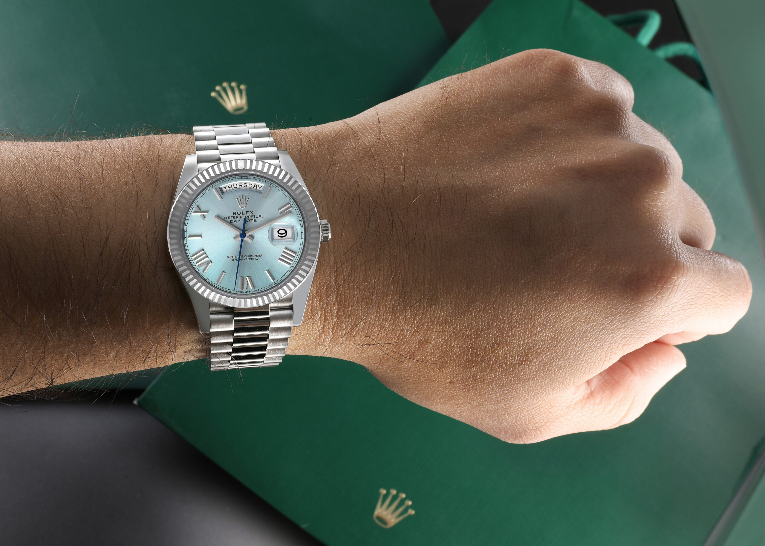 A platinum Rolex Day-Date with an ice blue dial worn on a wrist, positioned over a green Rolex box.