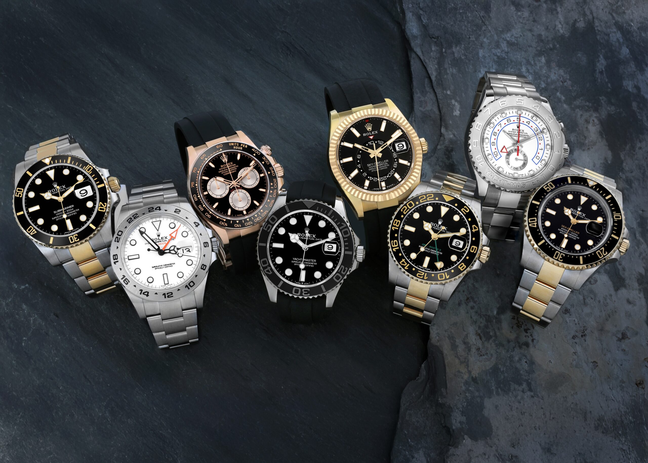 A group of professional Rolex watches, including a Submariner, GMT-Master II, and Yacht-Master, displaying both metal Oyster bracelets and black elastomer Oysterflex straps on a dark stone background.