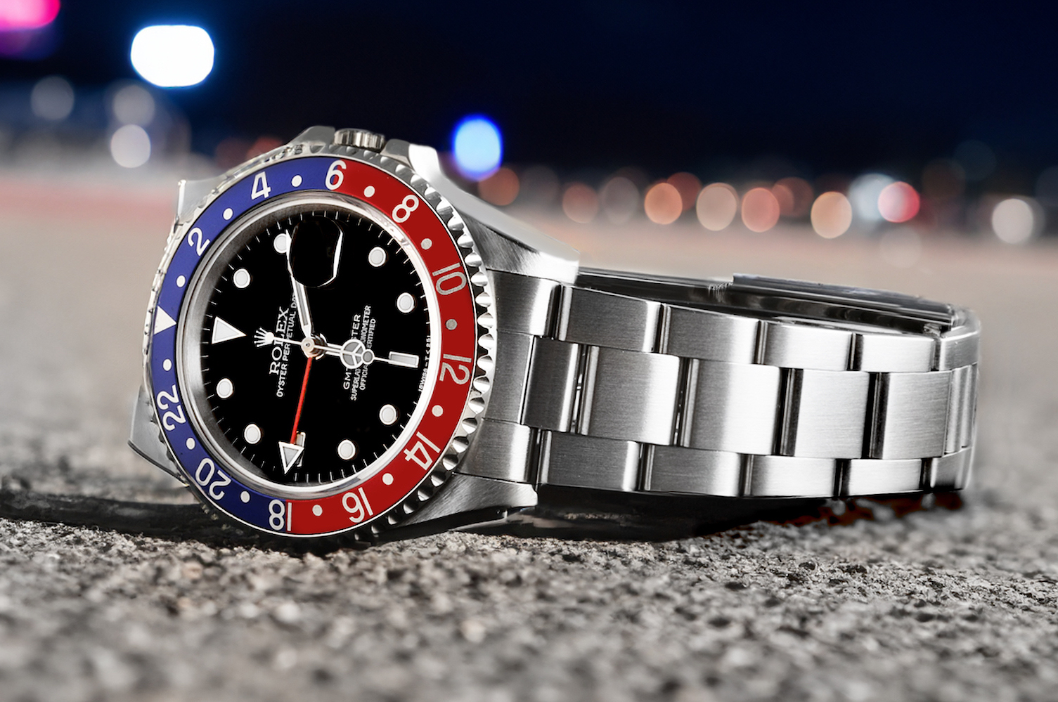 Rolex GMT-Master II 'Pepsi' (Ref. 16710) with a blue and red aluminum bezel, black dial, and stainless steel Oyster bracelet, with blurred city lights in the background.