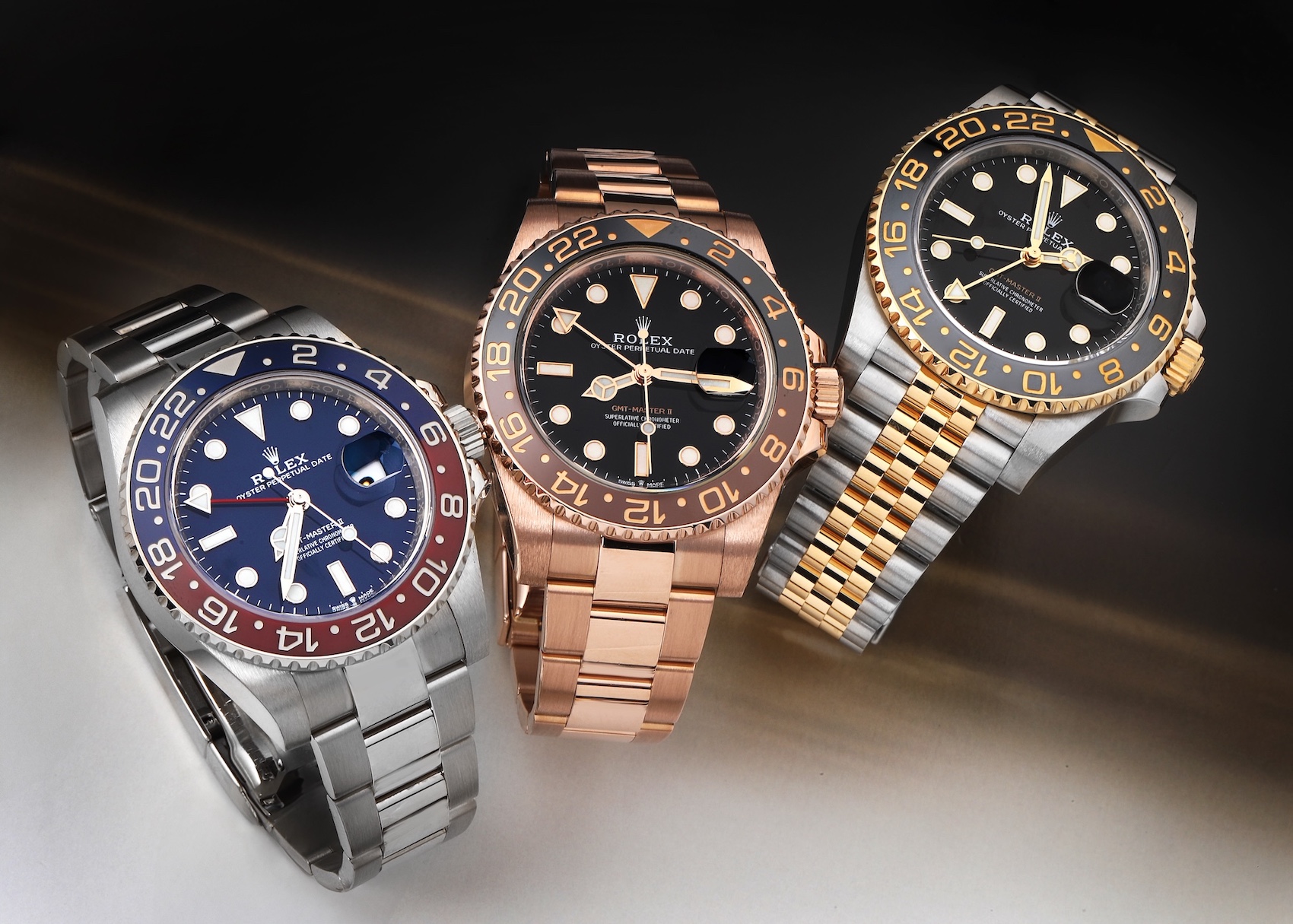 Three Rolex GMT-Master II watches are displayed on a gradient background: a stainless steel model with a blue and red "Pepsi" bezel and blue dial, an Everose gold model with a black and brown bezel and black dial, and a Rolesor (steel and yellow gold) model with a black and grey bezel and black dial on a Jubilee bracelet.