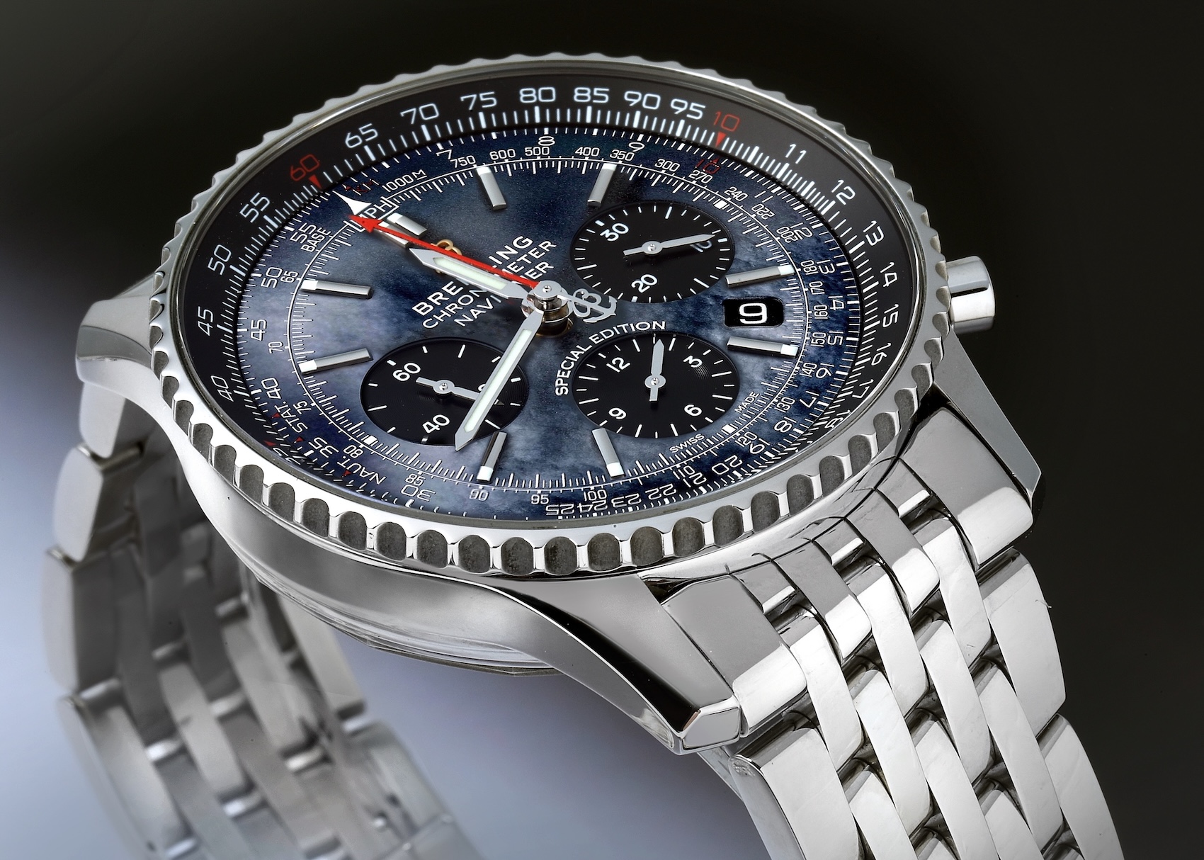 A detailed shot of a Breitling Navitimer 01 watch. It features a complex blue mother-of-pearl dial with three black subdials, a stainless steel case with a slide rule bezel, and a multi-link steel bracelet.