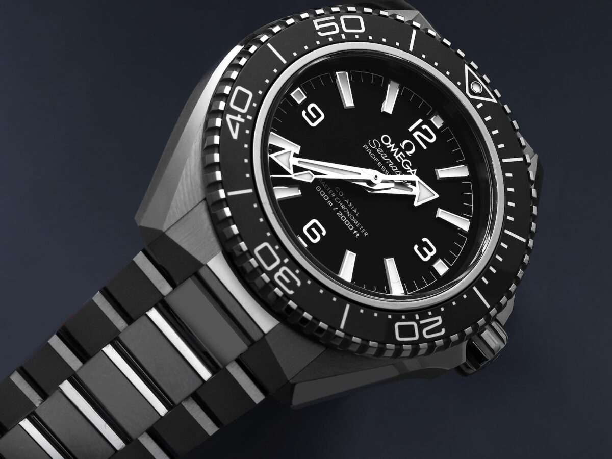 A dramatic, low-angle close-up photograph of the Omega Seamaster Planet Ocean 600M watch (ref. 217.30.42.21.01.001) set against a dark blue background. The shot highlights the sharp angular lines of the steel case, the deep black ceramic bezel, and the symmetrical no-date dial with open-work Arabic numerals. The watch is shown on an unconventional dark, reflective bracelet with polished center links.
