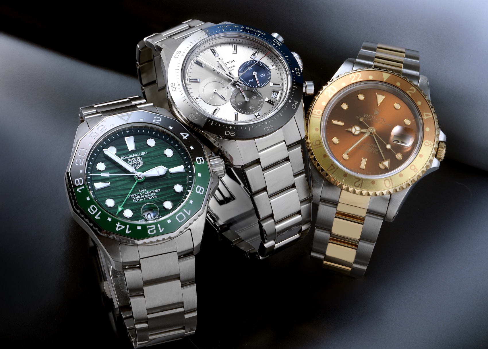 Group of three luxury mens watches featuring a green TAG Heuer Aquaracer, silver Zenith Chronomaster Sport, and two-tone Rolex GMT-Master II Root Beer.