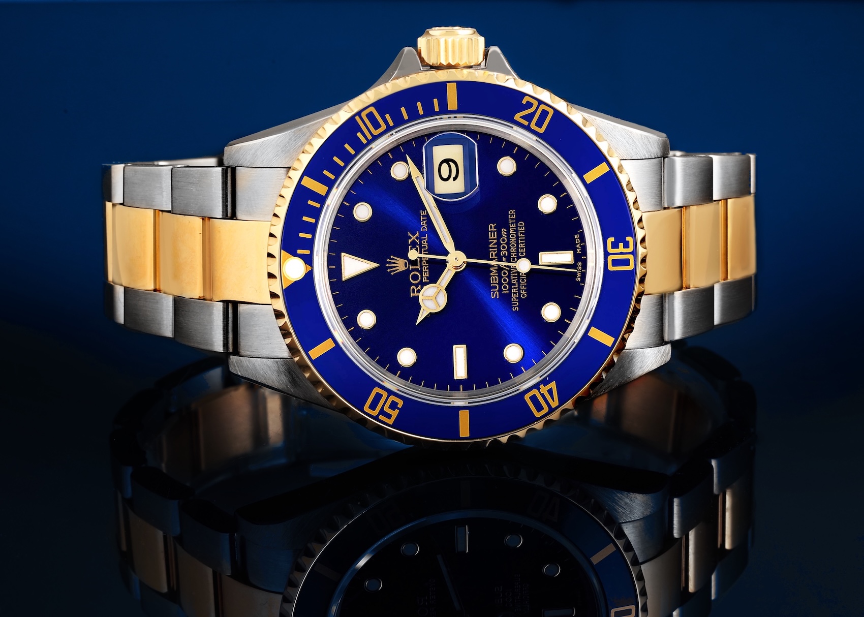 A sharp close-up studio photograph of a two-tone Rolex Submariner Reference 16613 ("Bluesy") with a cobalt blue sunburst dial and blue bezel, on a deeply reflective dark surface.