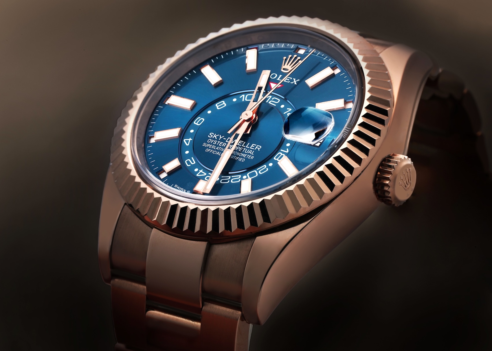  A close-up, dramatic angle photograph emphasizing the case profile and fluted bezel of a Rolex Sky-Dweller in Everose Gold. The dynamic teal tones of the blue-green dial are visible as they shift in the warm light, paired with an Oyster bracelet.